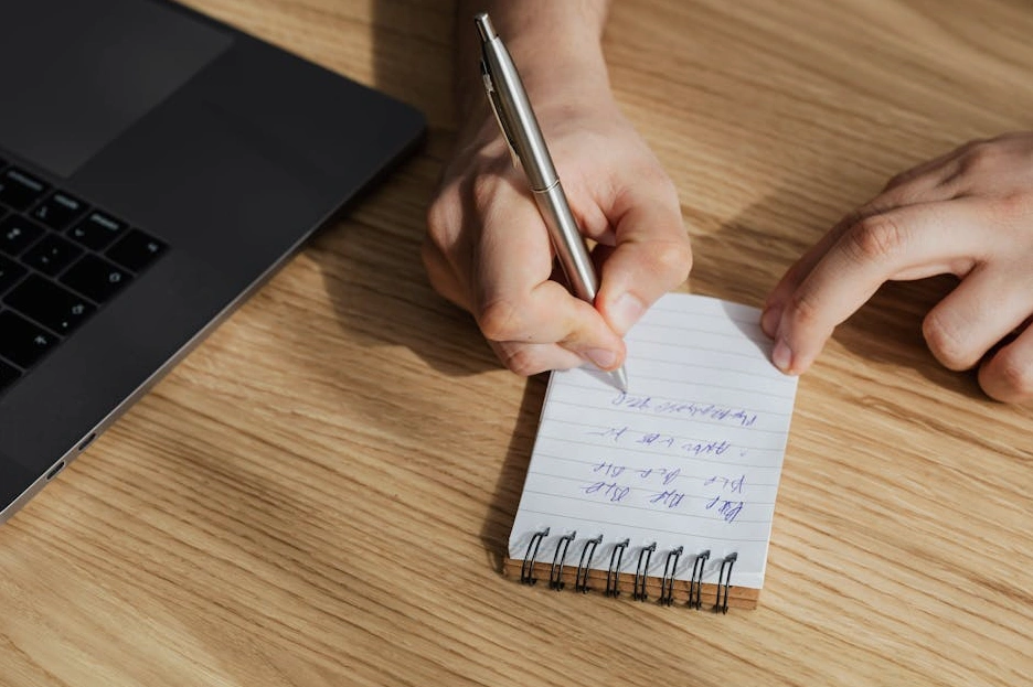 Osoba si zapisuje poznámky do sešitu u notebooku